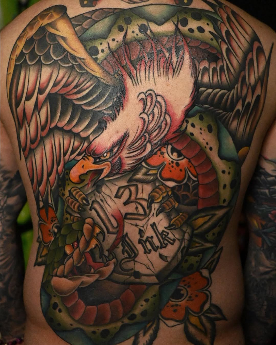 Colorful back tattoo featuring an eagle with spread wings, snake coiled around the body, fish, and script lettering in red, green, orange, and gold tones.