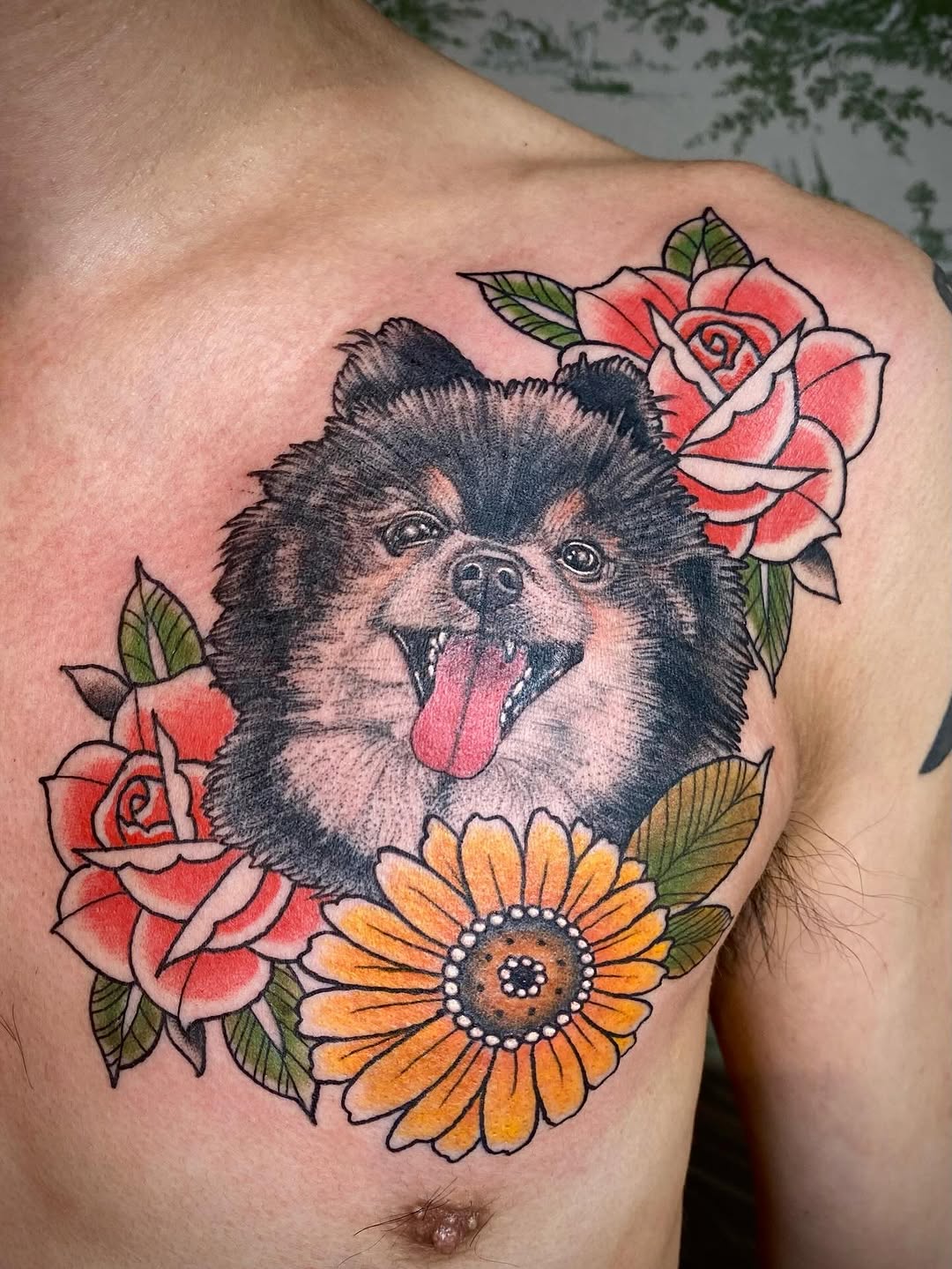 A smiling small dog with pink tongue surrounded by red roses and a yellow sunflower with green leaves, tattooed on the chest.