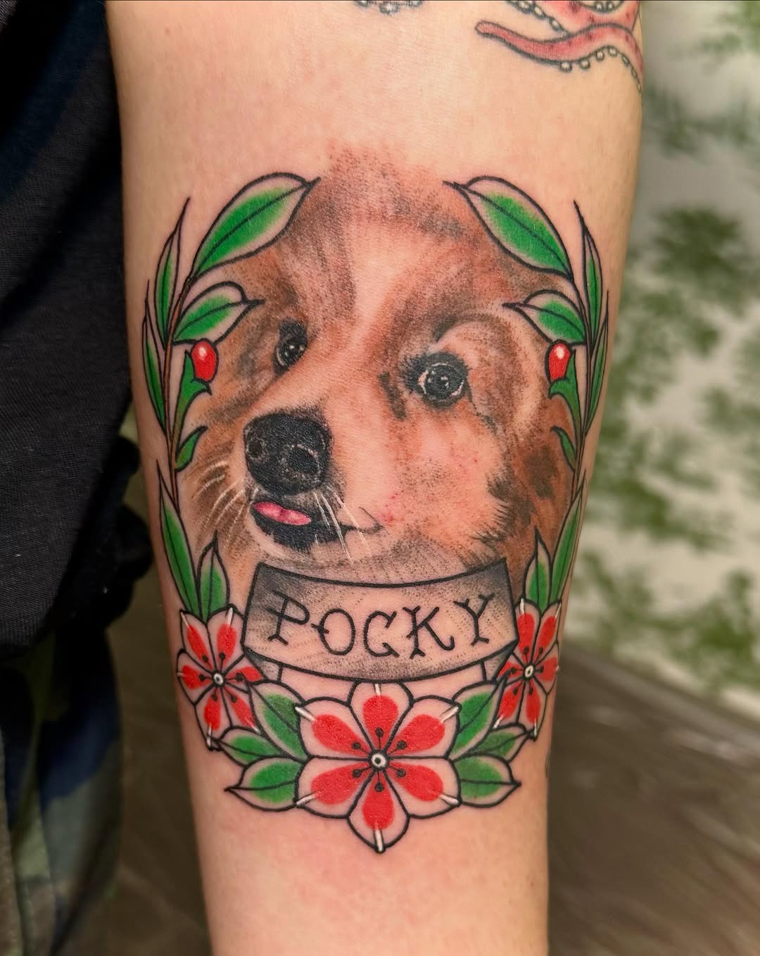 Portrait tattoo of a brown dog named Pocky with a pink tongue, framed by red flowers and green leaves on the upper arm.