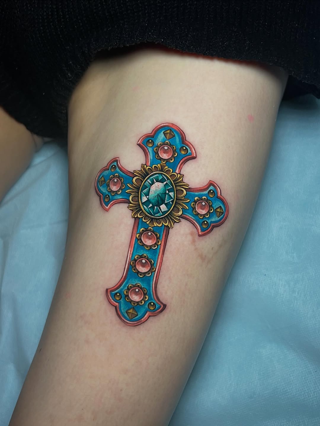Ornate cross tattoo on upper arm featuring a turquoise body with red outline, coral gemstones, gold floral centerpiece, and teal faceted stone at center.