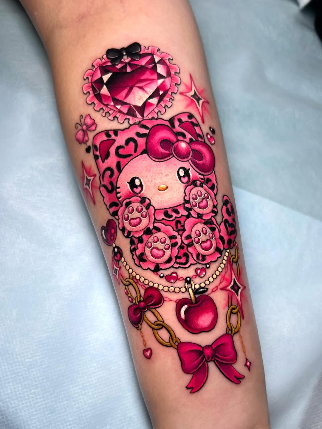 Hello Kitty character tattoo on forearm featuring a pink jeweled heart at top, the character's face with leopard print, paw prints, gold chain, red apple pendant, and red bows in shades of pink and red.