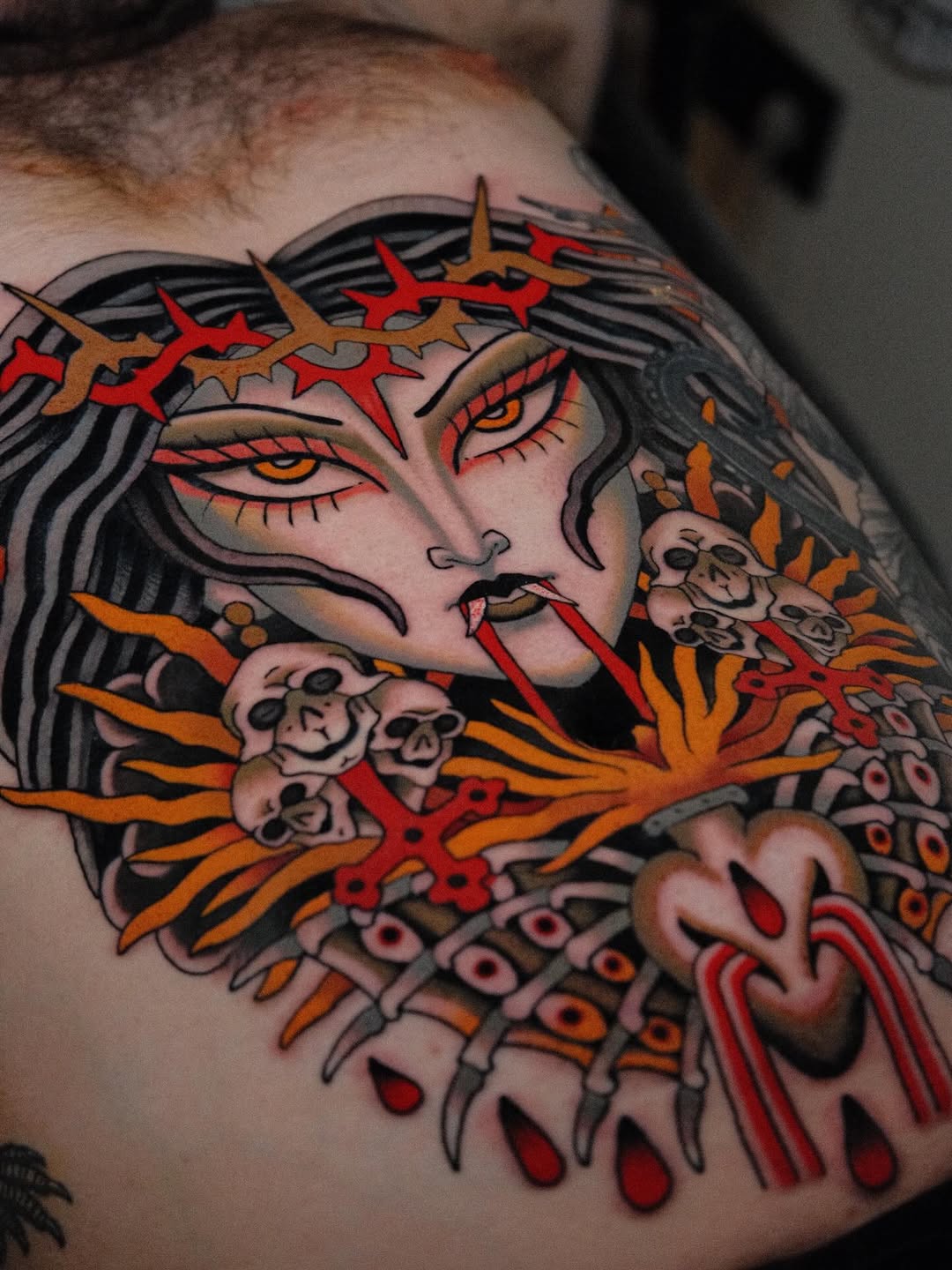 Colorful shoulder tattoo featuring a face with crown of spikes, closed eyes with red makeup, skulls, and orange and red wing-like elements.