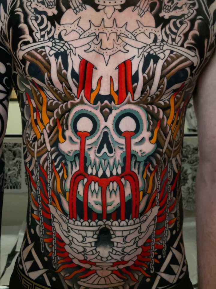Large chest and torso tattoo featuring a skull with red eyes and mouth, surrounded by flames, chains, and ornamental designs in red, black, and gold.