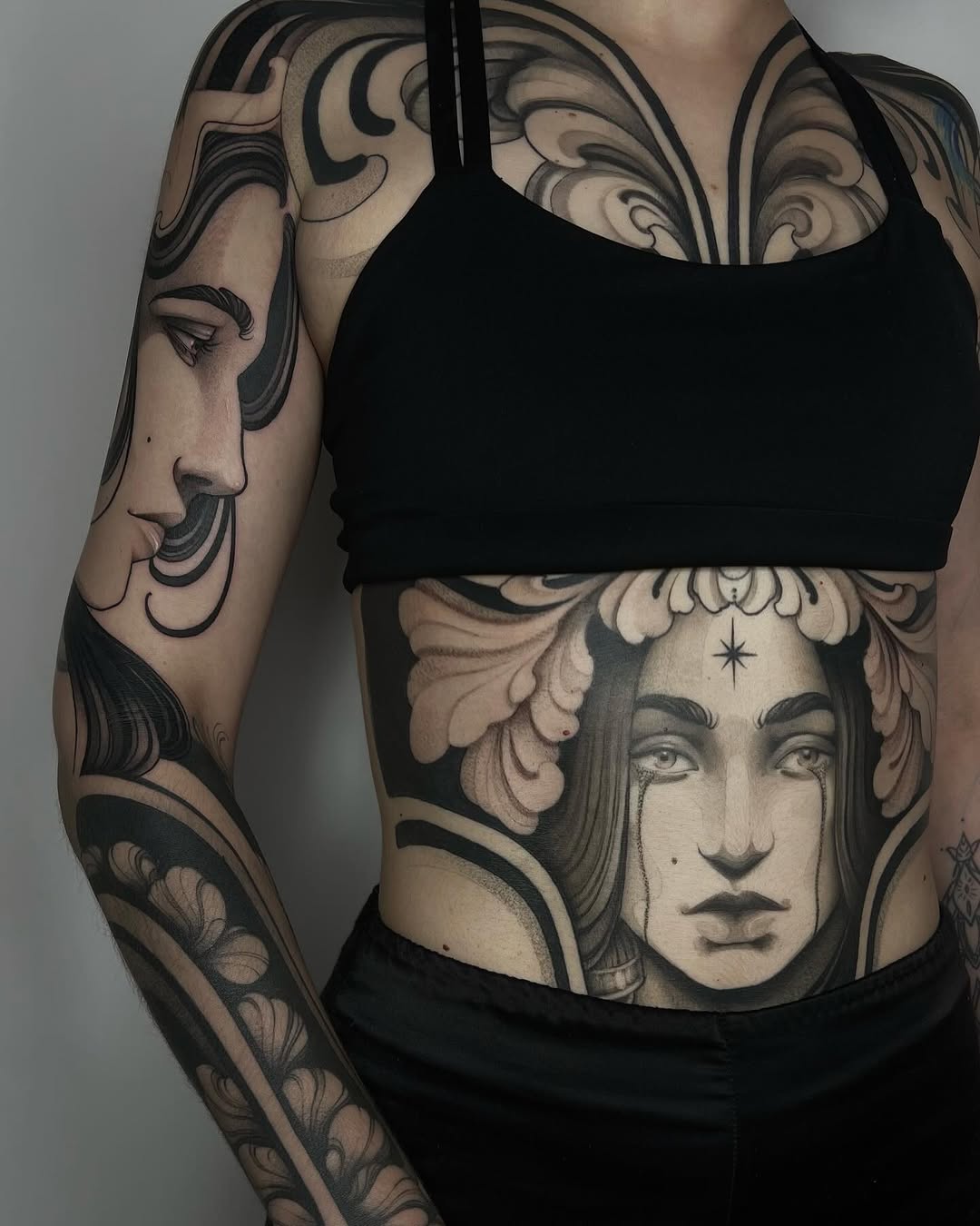 Detailed portrait tattoos on arm and torso featuring two faces with ornamental flourishes and decorative elements in black and grey ink.