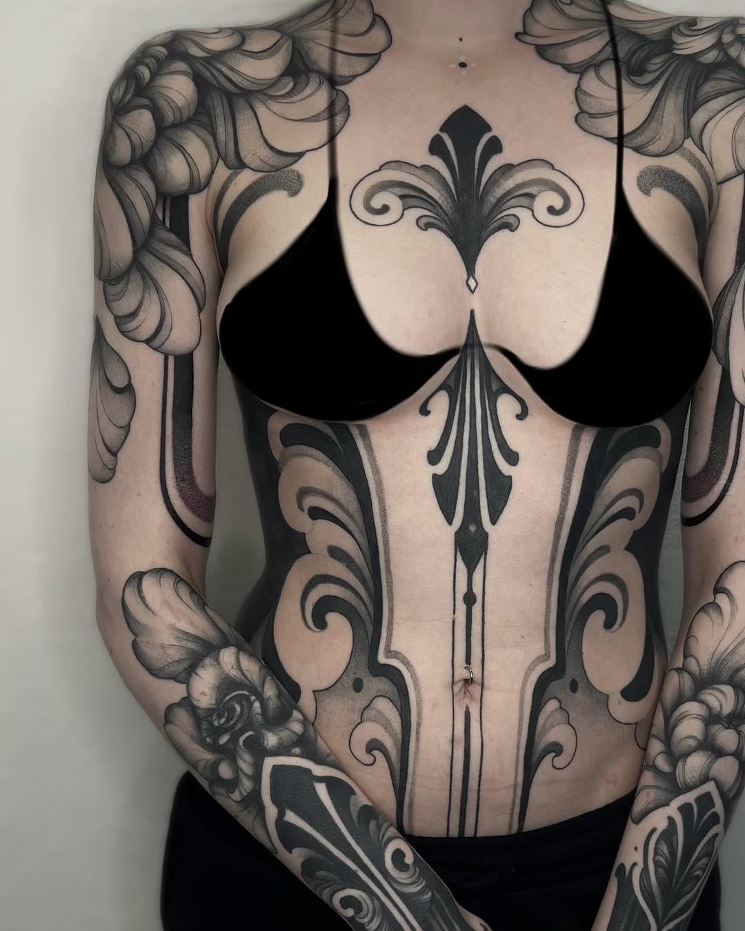 Ornate symmetrical chest and arm tattoos featuring scrolling flourishes, fleur-de-lis motifs, and decorative patterns in black ink on pale skin.