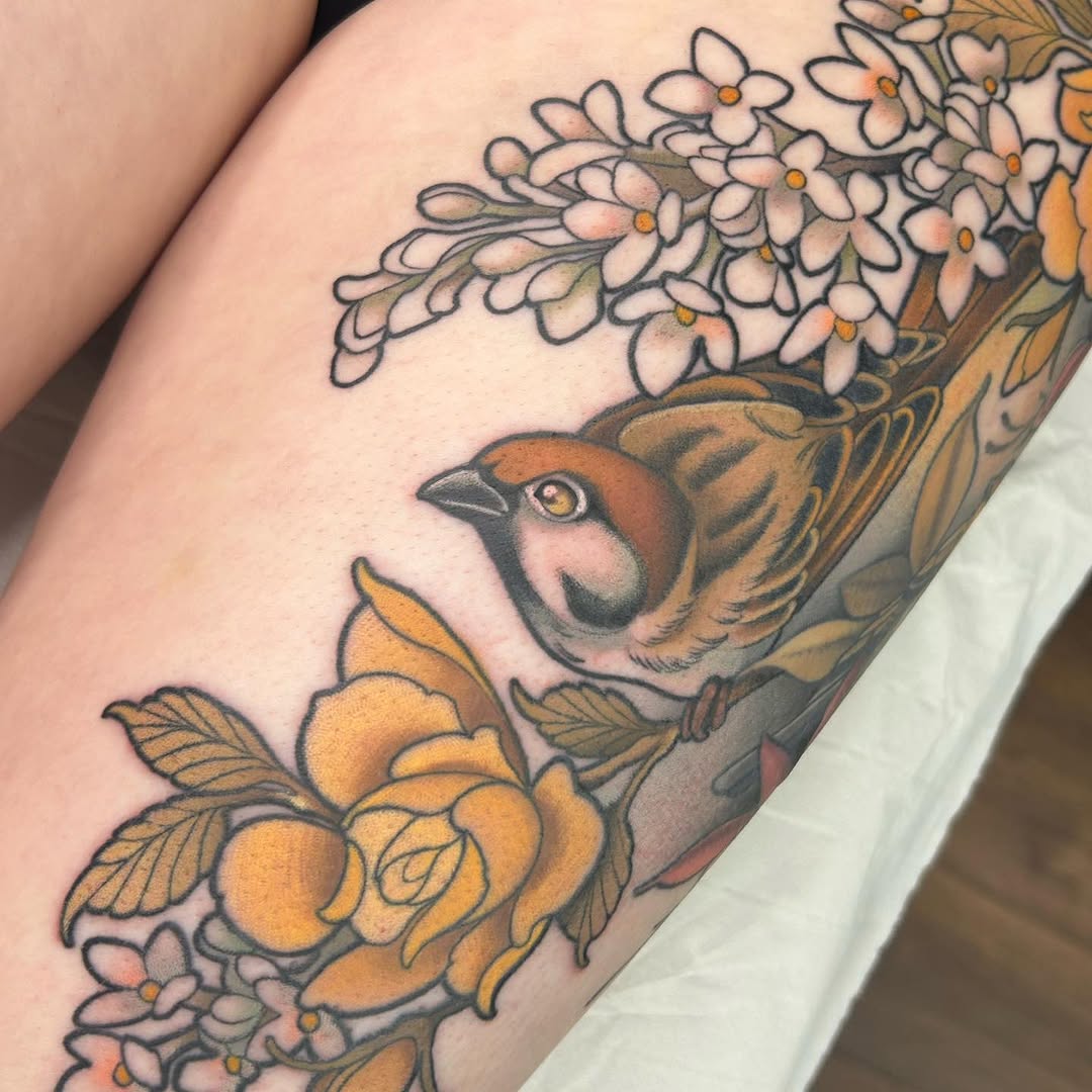 Colorful tattoo on an arm featuring a brown bird with yellow eye surrounded by white flowers with yellow centers and yellow fruits with green leaves.