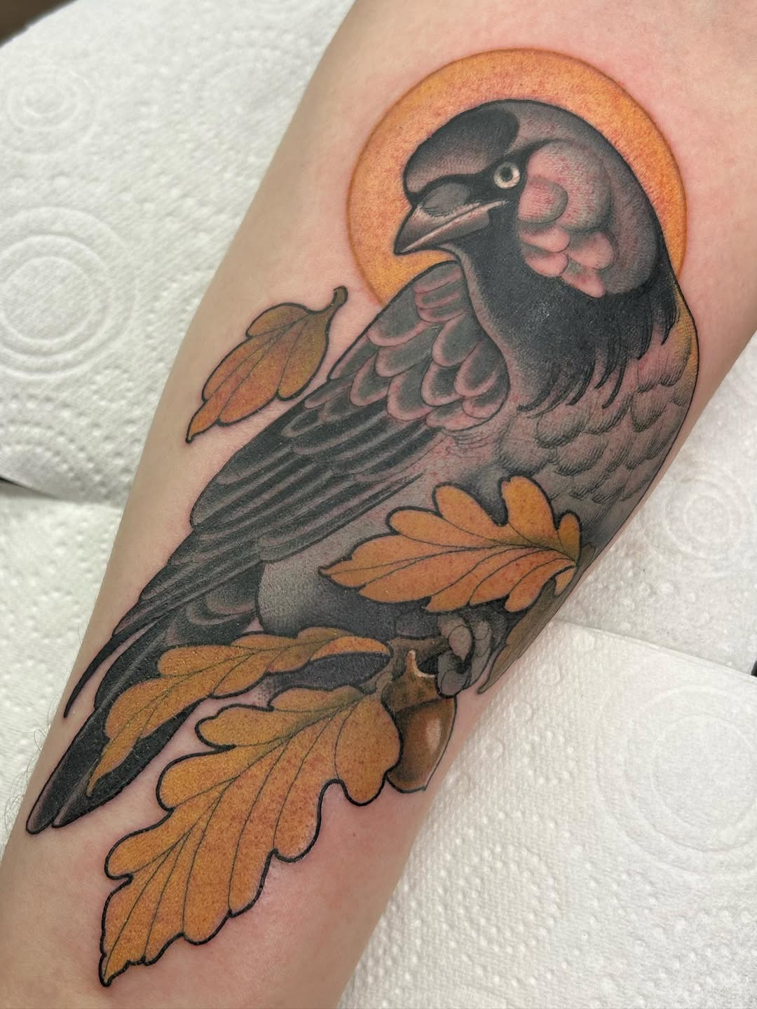 A dark bird with a golden halo and pink wing accents perched on brown oak leaves, tattooed on the upper arm.