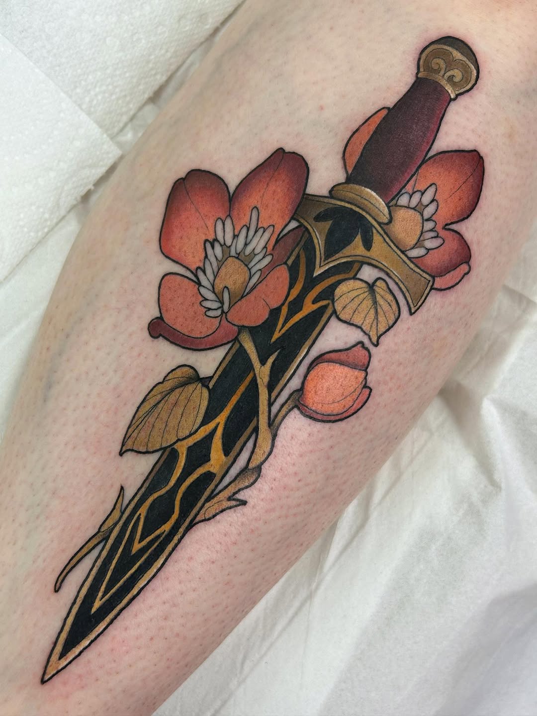 Decorative dagger with ornate gold hilt and burgundy wrapping, adorned with coral-red flowers and gold leaves, tattooed on the upper leg.