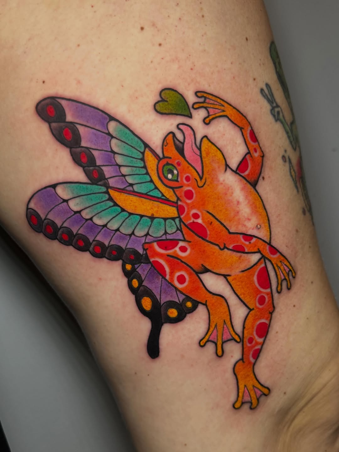 Colorful mythical creature tattoo on thigh featuring purple and teal wings with red spots, orange body with red patterning, and green leaf detail.