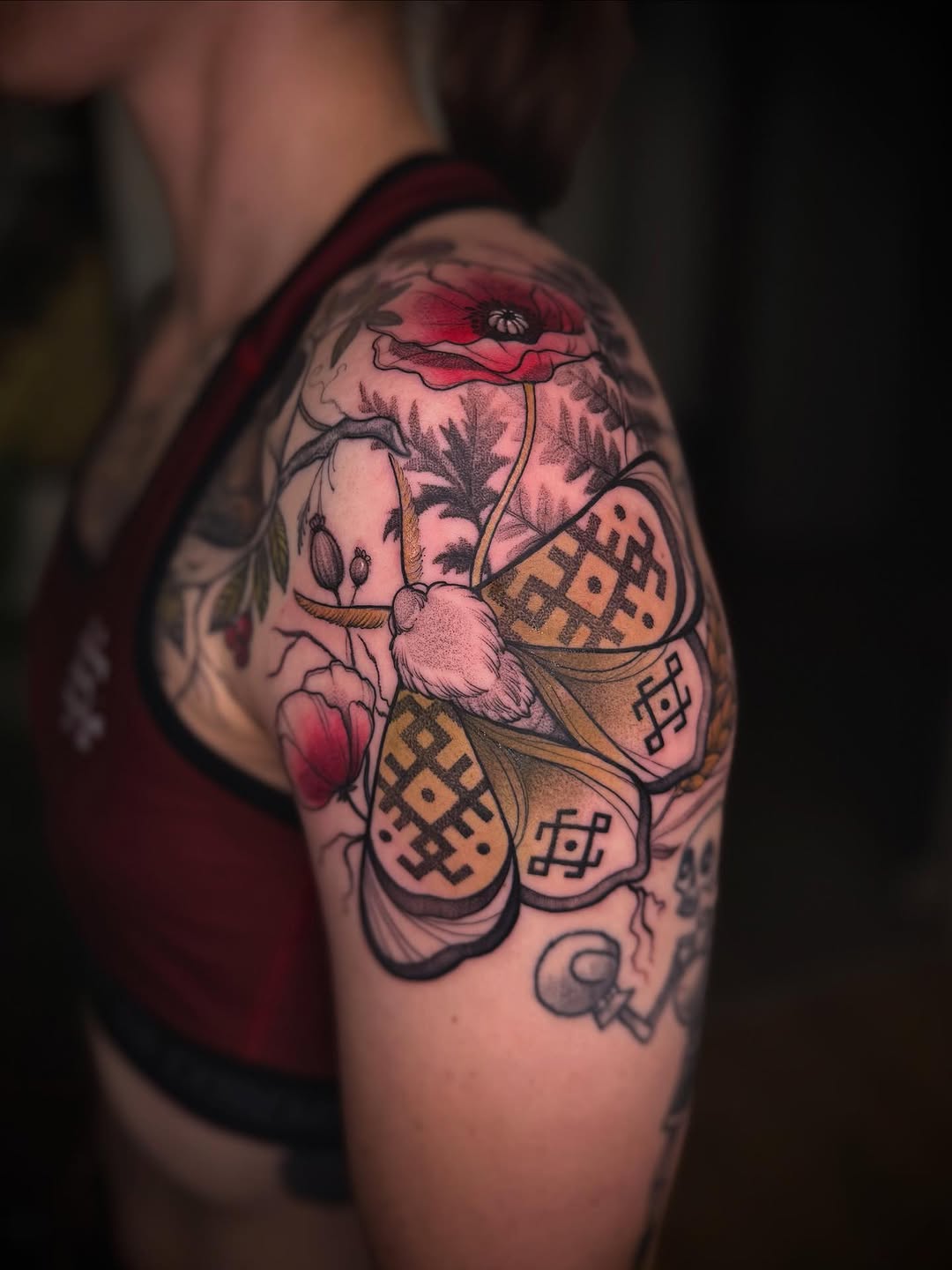 Colorful shoulder tattoo featuring red poppies, a butterfly with patterned wings, botanical elements, and geometric designs in red, gold, and black ink.