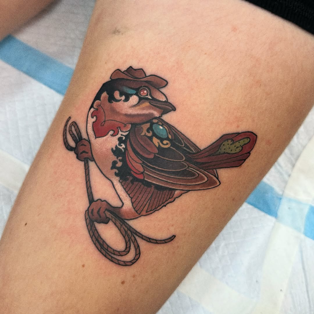 Cowboy bird character tattoo on forearm with brown, red, and teal colors, holding a lasso and wearing a hat.