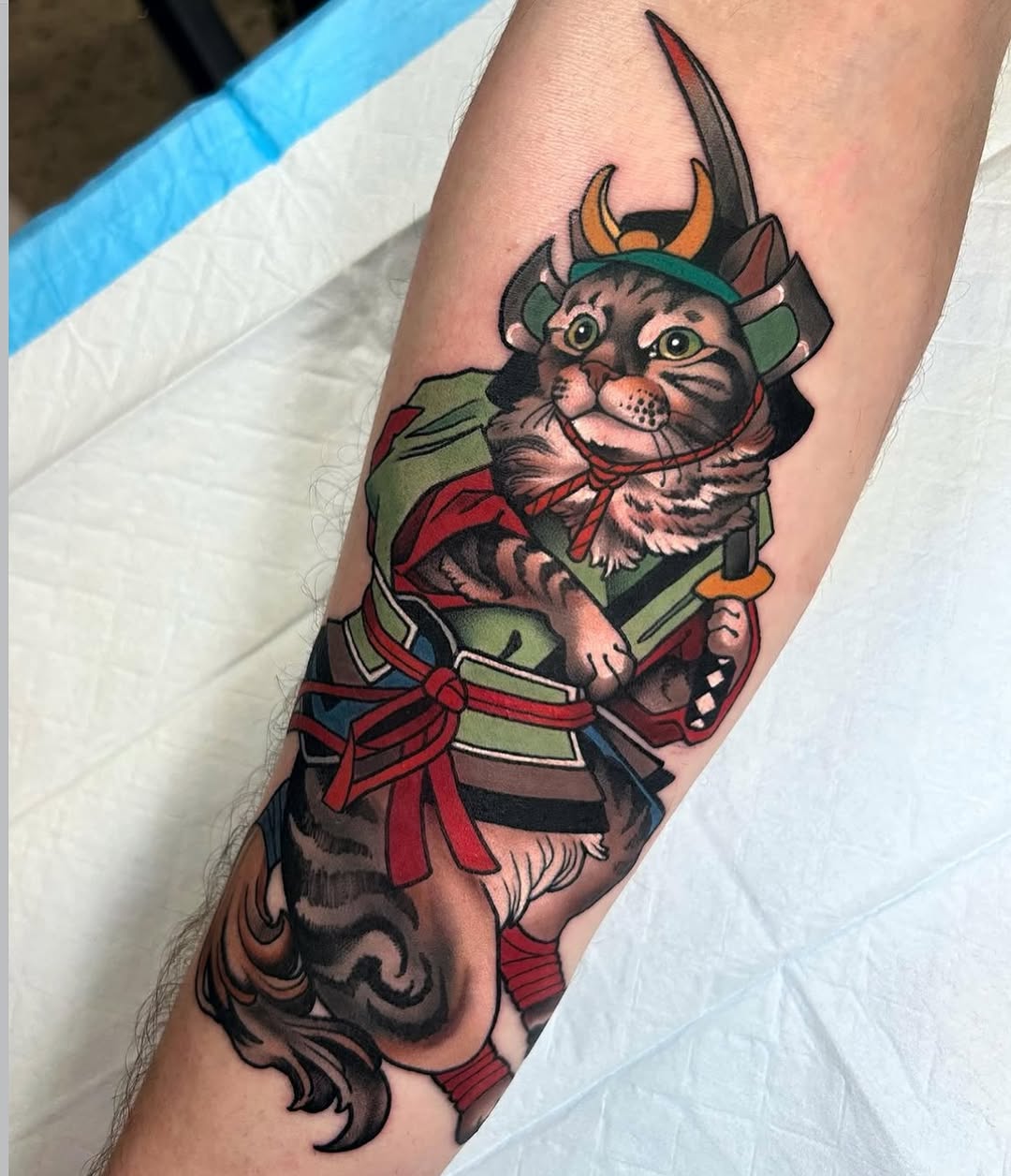 Colorful cat demon character with horns and sword tattoo on forearm, featuring green, red, and orange tones with decorative tail design below.