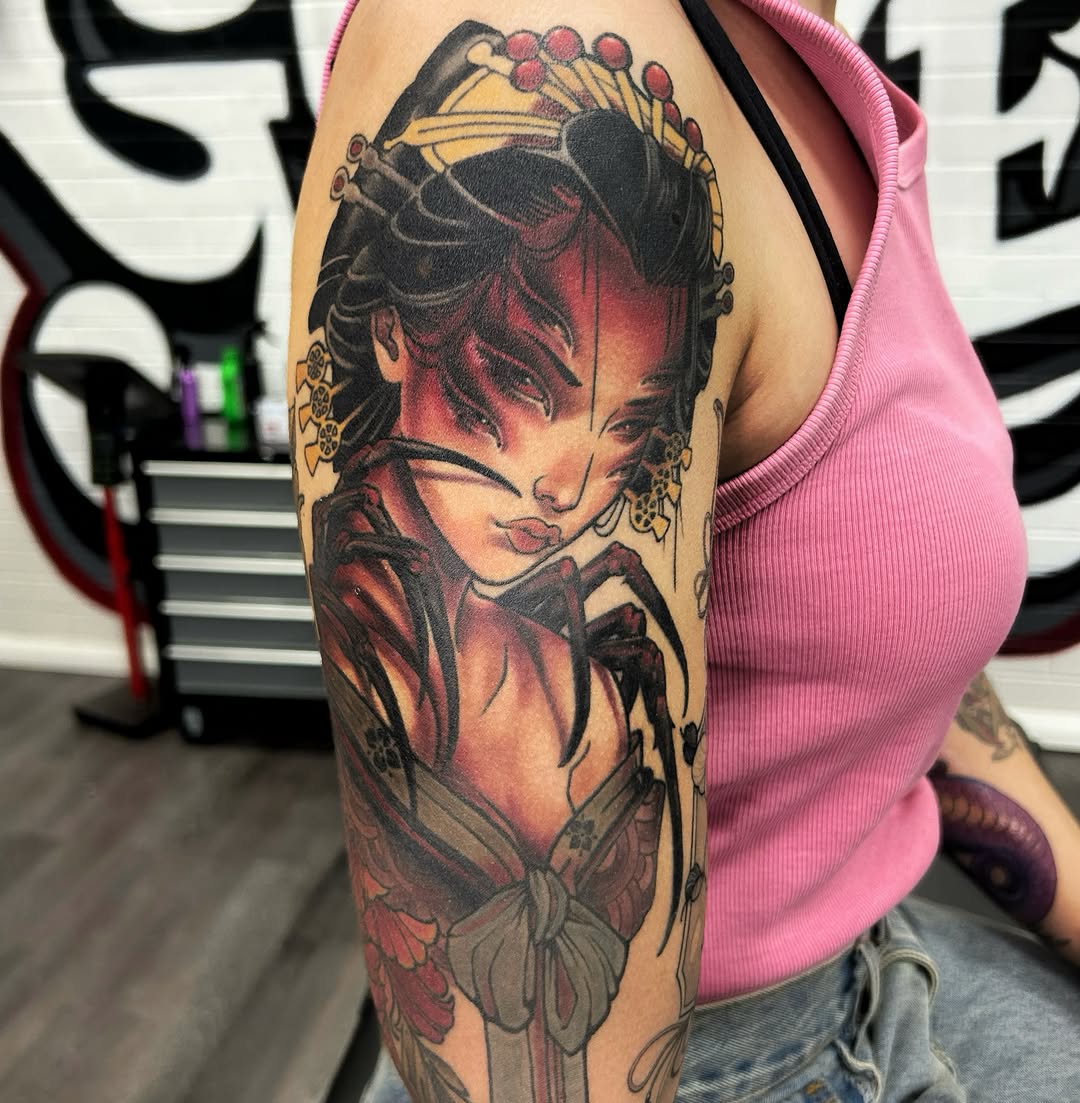 Colorful shoulder tattoo featuring a woman's face with closed eyes, decorative elements, and floral motifs in red, black, and gold tones.
