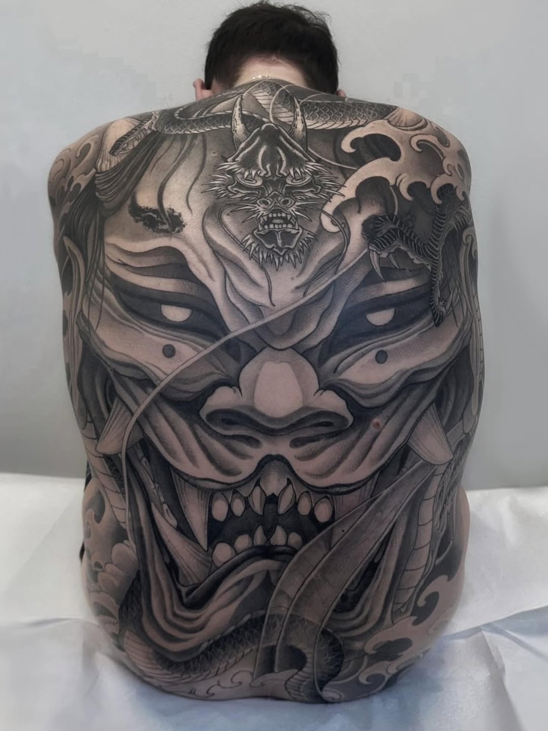 Large-scale back tattoo featuring a demonic mask with horns, fangs, and ornate decorative elements in black and grey ink.