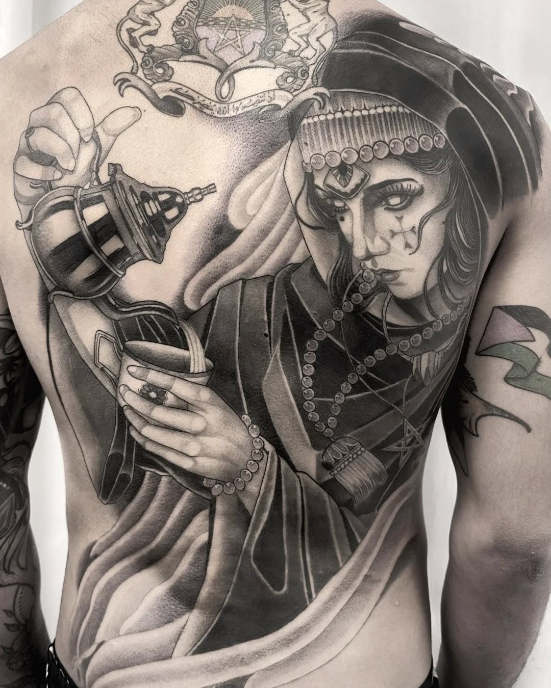 Large-scale back tattoo featuring an ornate female figure in elaborate headdress and jewelry holding a decorative sphere, with heraldic crest above, rendered in grayscale ink.