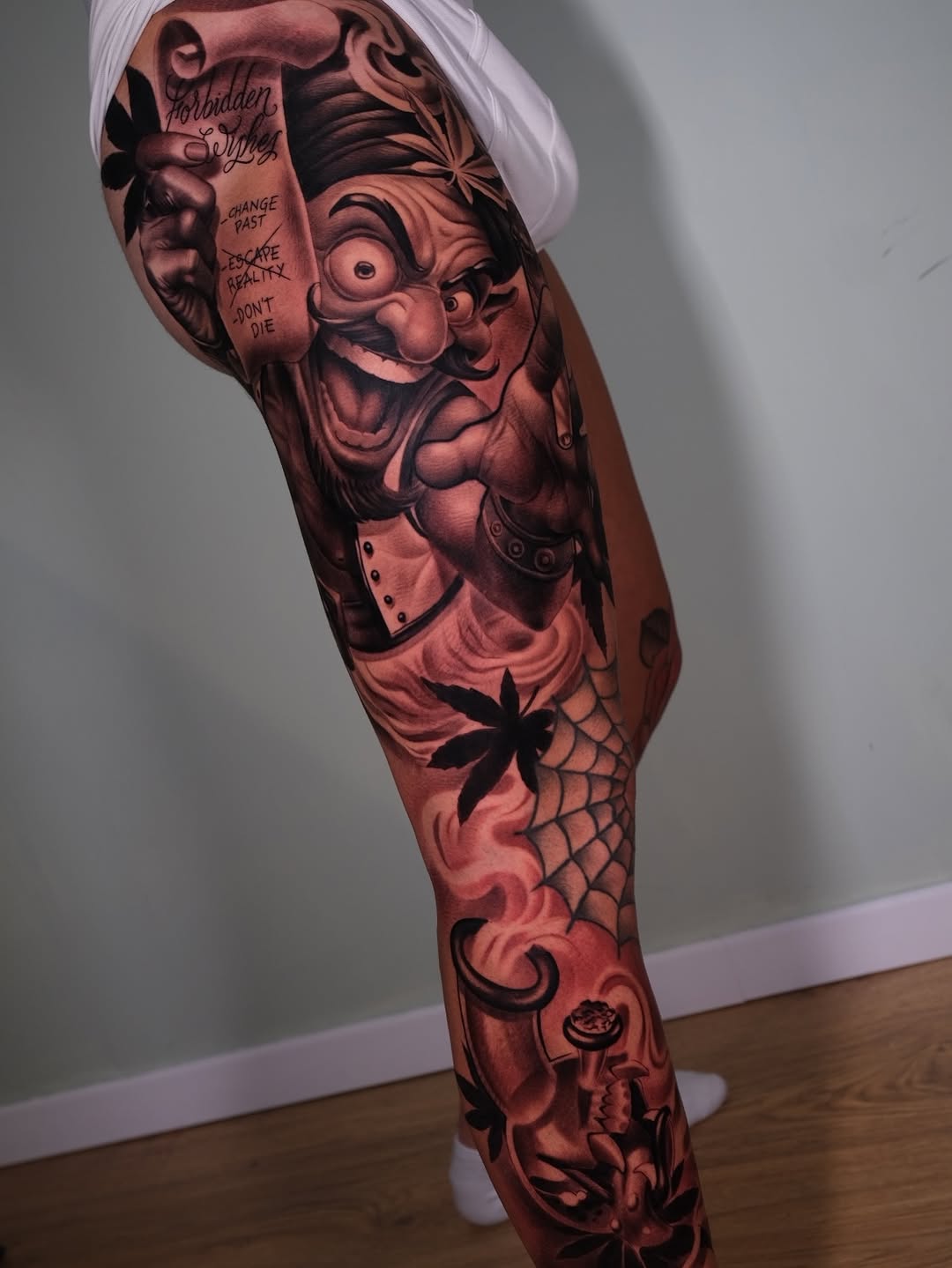 Full leg sleeve tattoo featuring a laughing jester character with text, flowers, spider web, and decorative swirls in red and black ink.