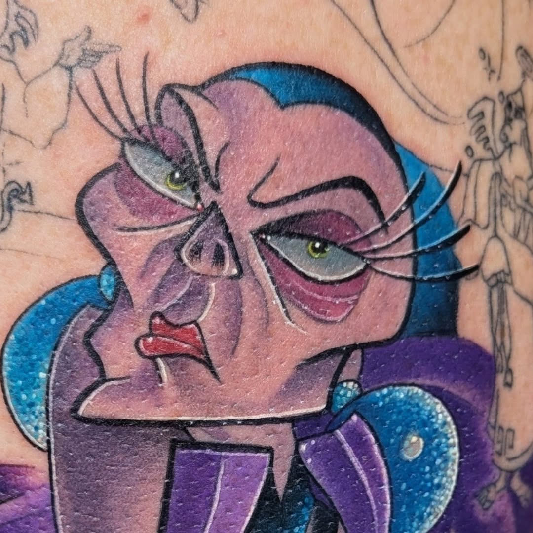 Colorful shoulder tattoo featuring a stylized female face with purple skin, yellow eyes, long lashes, red lips, and blue hair against skin.