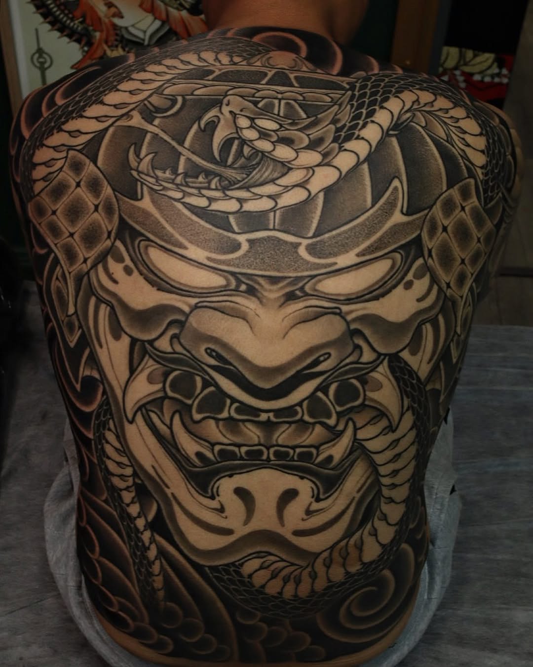 Large back tattoo featuring a multi-faced demon mask design with intricate scale patterns, shading, and ornamental details in black and tan ink.