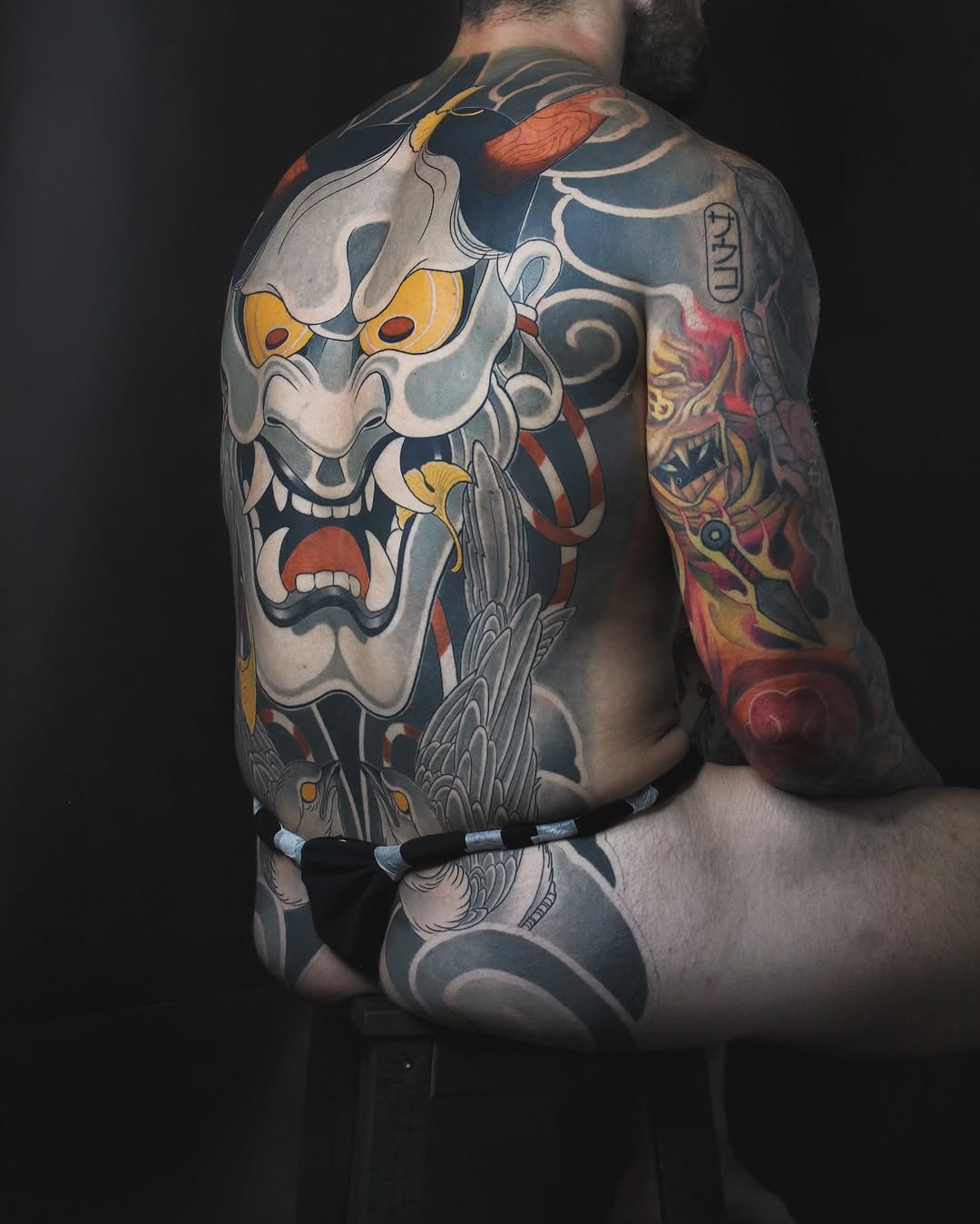 Back tattoo featuring two large demon masks with gold and red accents, ornamental details, and a red arm piece with decorative motifs on dark skin.
