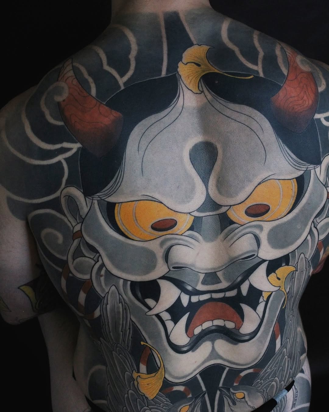 Large back tattoo featuring a white and grey demon mask with gold accents, red mouth, and ornamental designs on dark background.