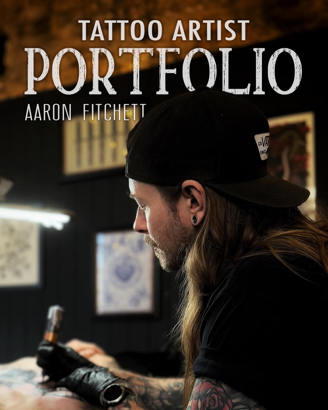 Tattoo artist Aaron Fitchett wearing a black cap and long-sleeved shirt displays an arm tattoo in red and dark colors while working in his studio.