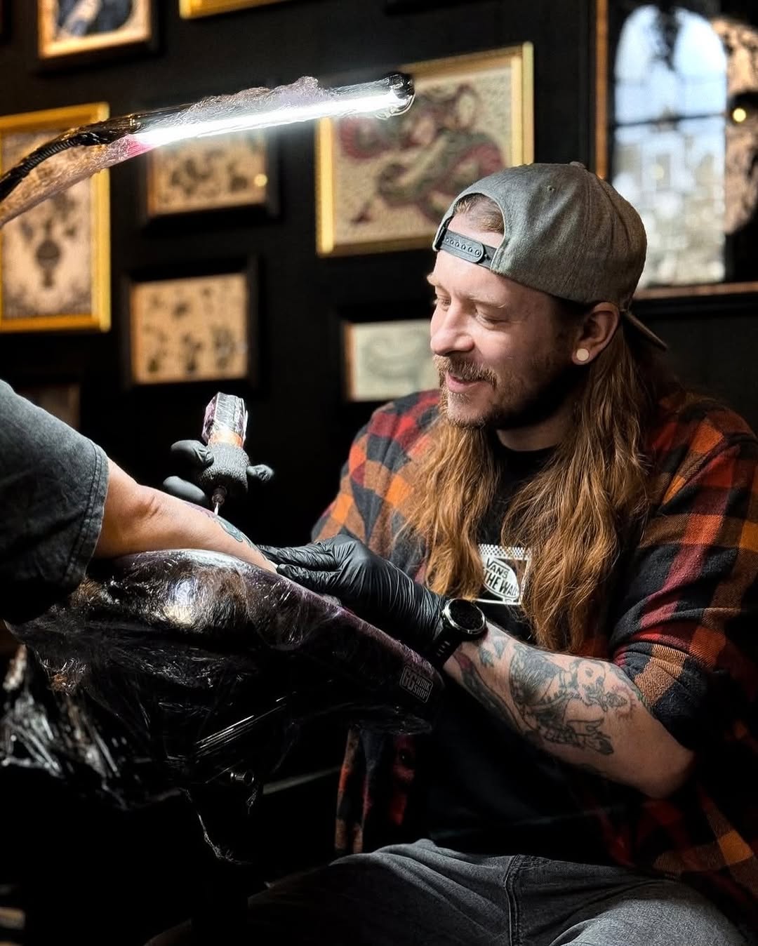 Tattoo artist with long hair and beard wearing a plaid shirt examines a large dark floral design on a client's arm under bright lighting.