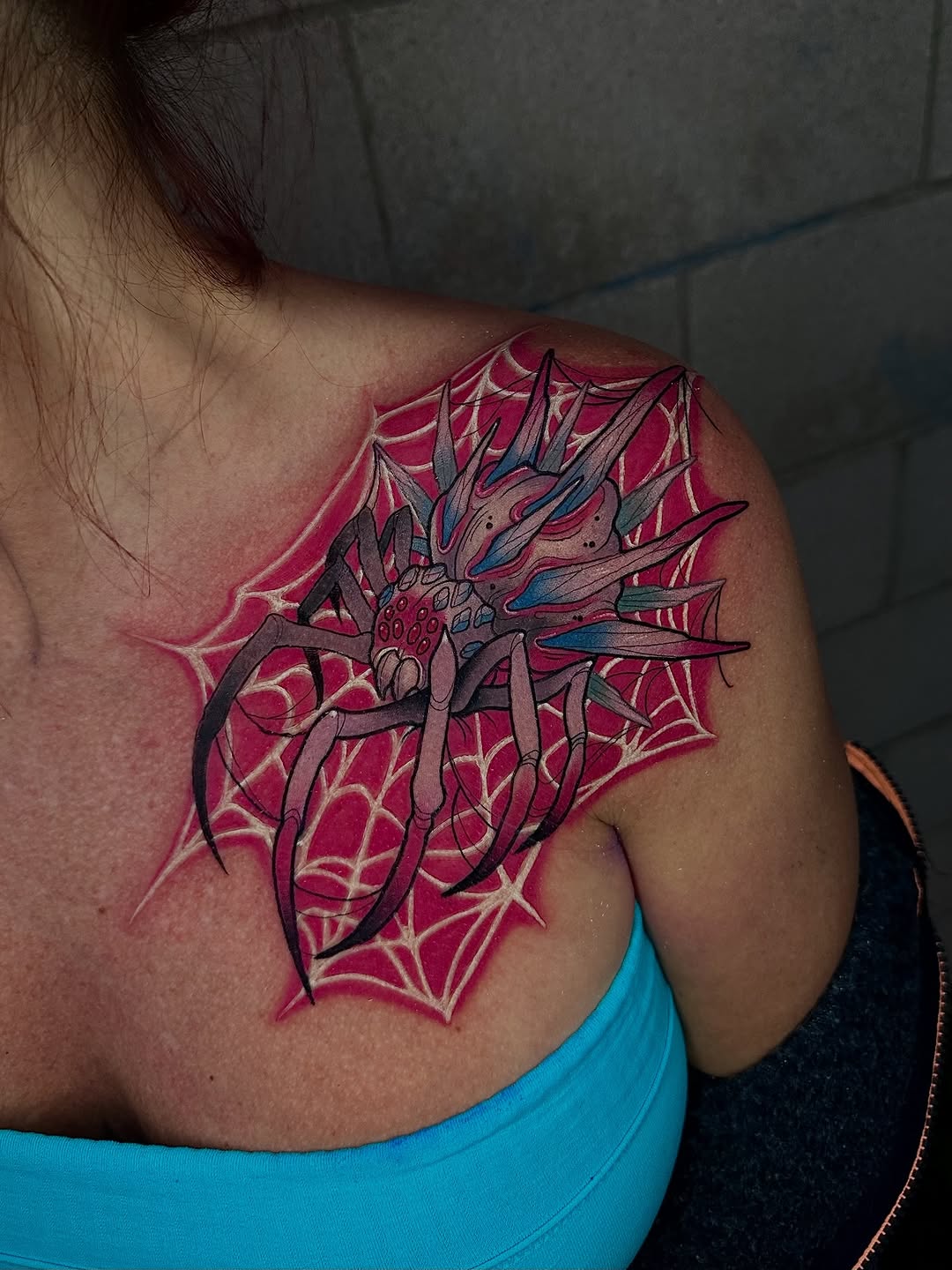 Colorful spider with spiky legs positioned on shoulder blade, featuring magenta and pink web background with blue and gray details.