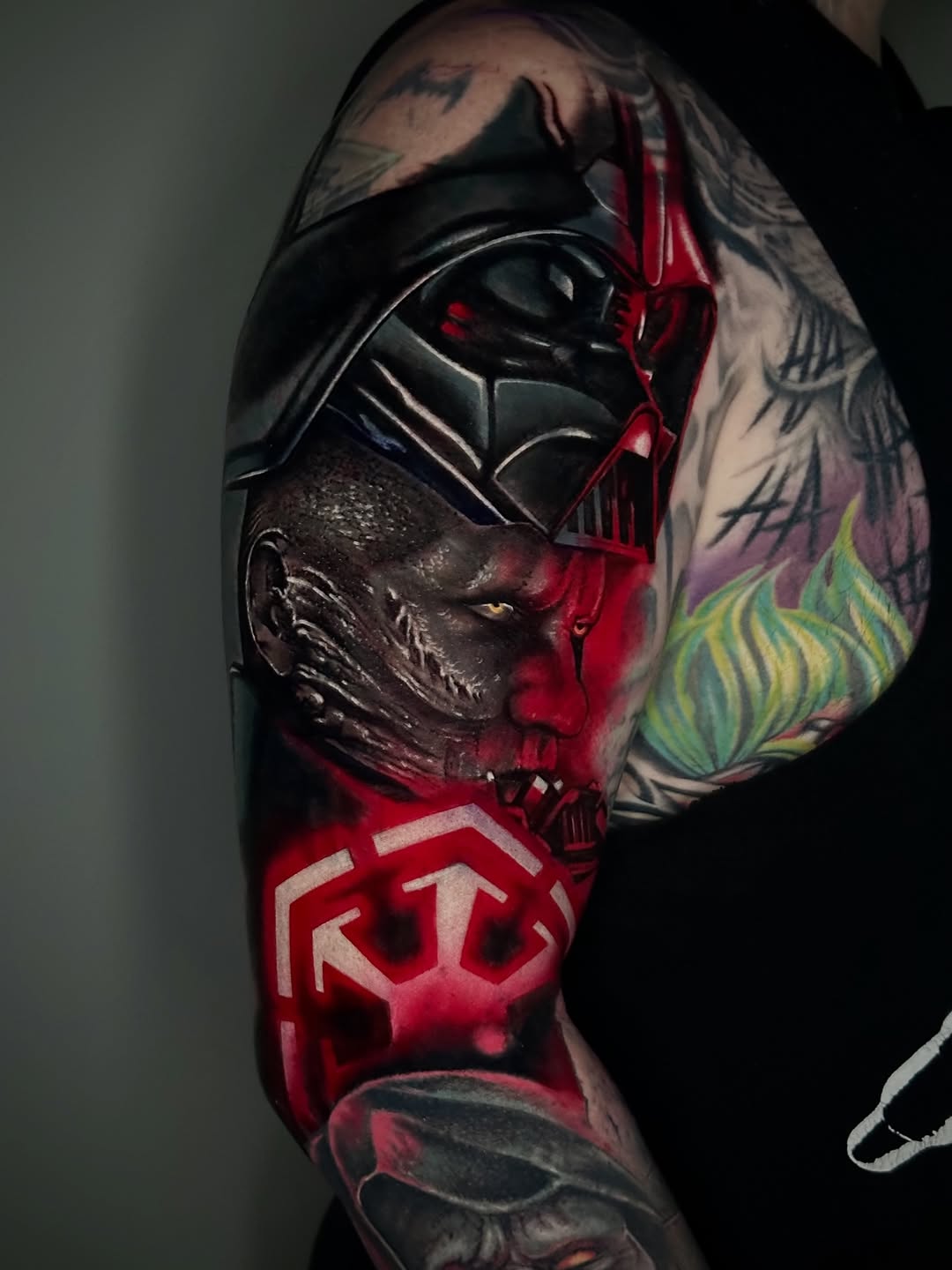 Colorful shoulder and arm tattoo featuring Darth Vader's helmet, a skull, red symbols, green and yellow flowers, and various dark imagery.