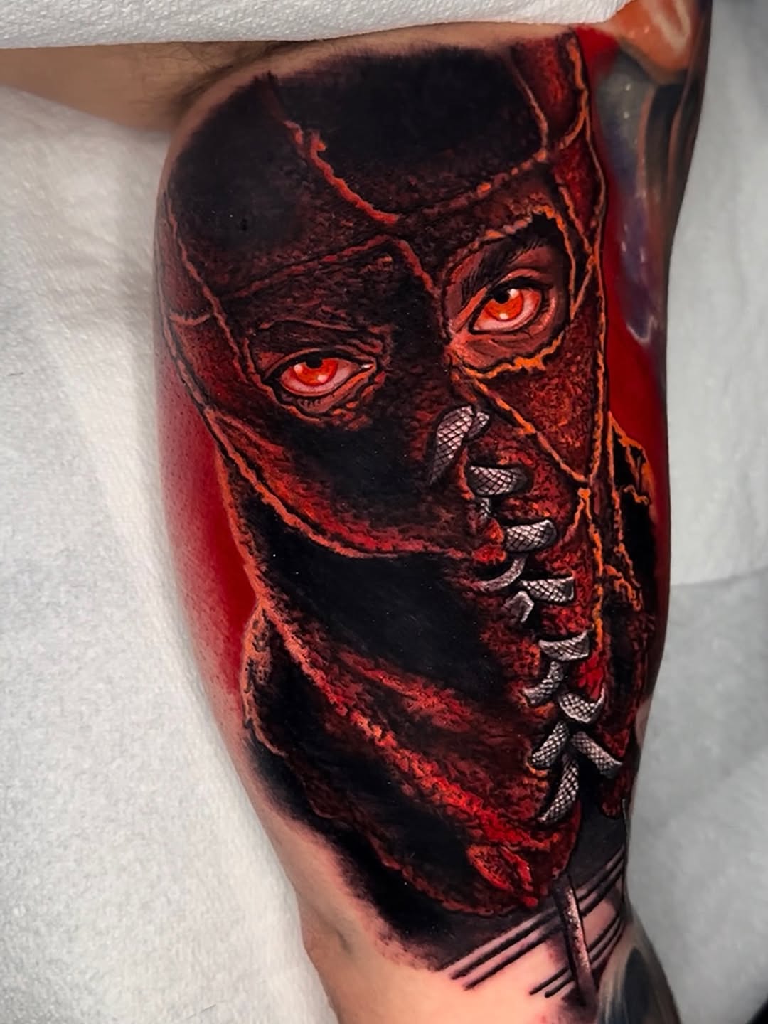 Demonic masked face with glowing red eyes and exposed teeth tattoo on upper arm in red, orange, and black ink.