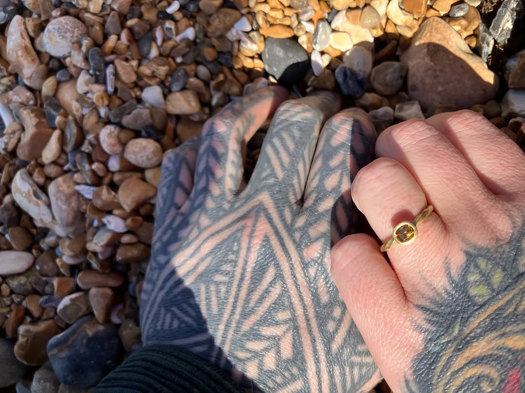 Detailed geometric and leaf pattern tattoo covering the hand and fingers in black and grey ink, displayed on pebbled beach background.