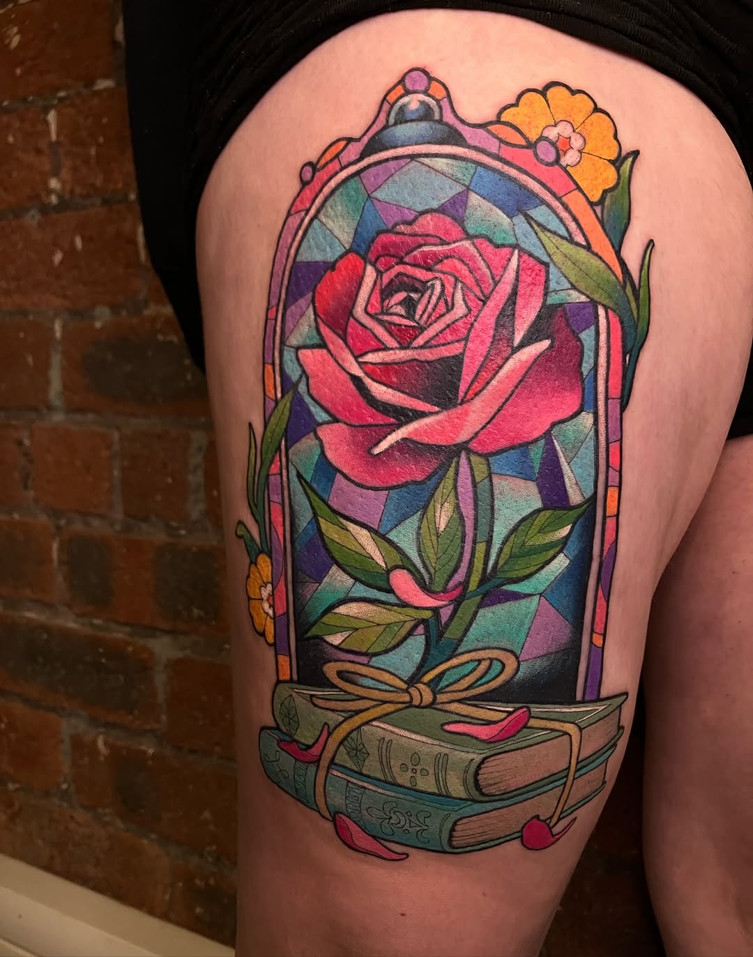 Colorful shoulder tattoo featuring a red rose in a glass dome with stained glass background, flanked by small flowers and stacked books with a bow.
