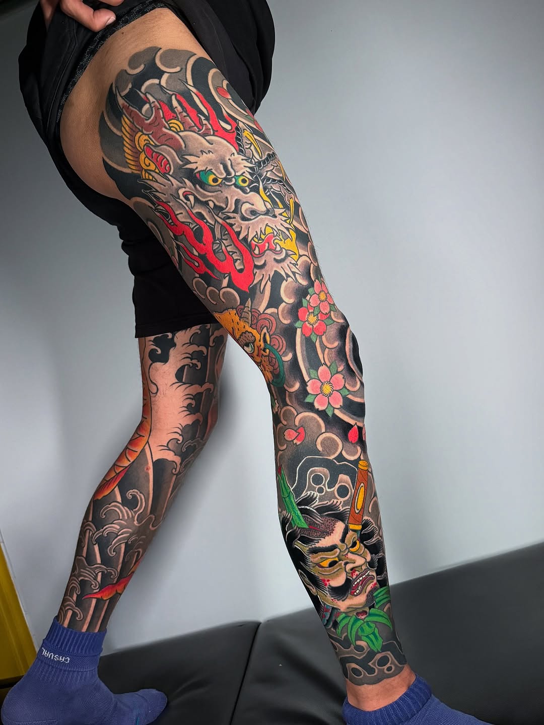 Full-leg tattoo sleeve featuring mythical creatures, ornamental scrollwork, red flowers, and colorful dragon and demon imagery in vibrant reds, yellows, greens, and blacks.