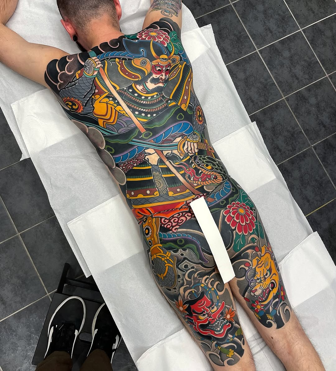 Full-leg color tattoo featuring a samurai warrior with ornate armor, demons, flowers, and decorative patterns in gold, red, blue, and green on dark background.