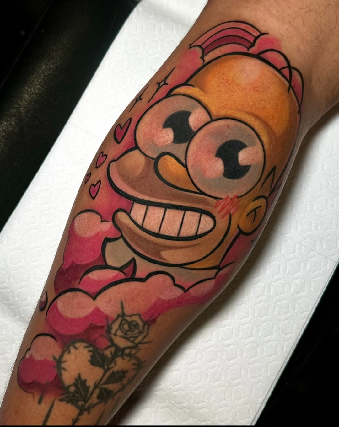 Colorful cartoon character tattoo on forearm featuring a smiling face with large eyes, surrounded by red hearts, clouds, and a rose, rendered in orange, red, and brown tones.
