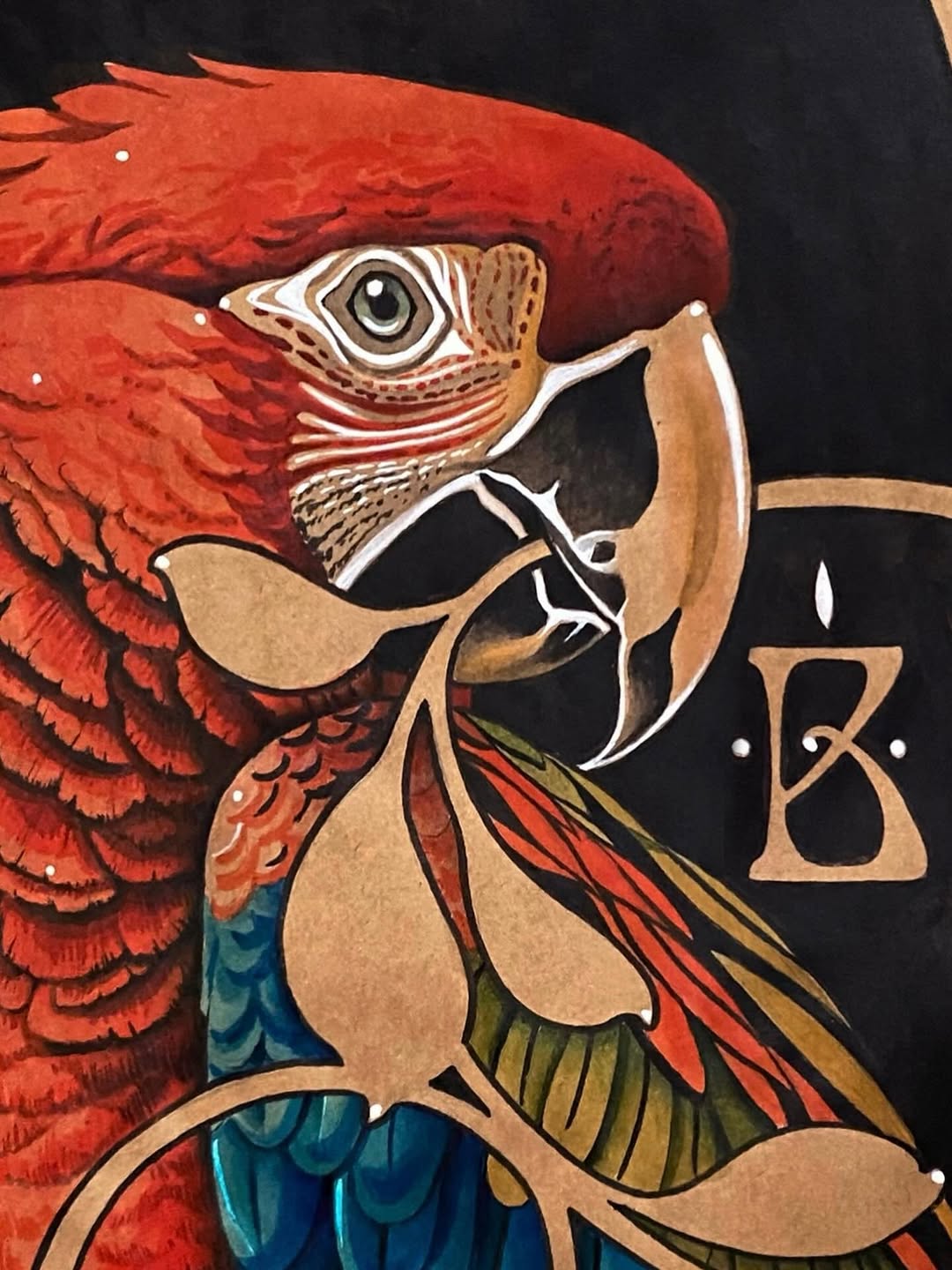 Vibrant red macaw head tattoo with detailed feathering, tan and blue wing sections, and a decorative letter B on black background.