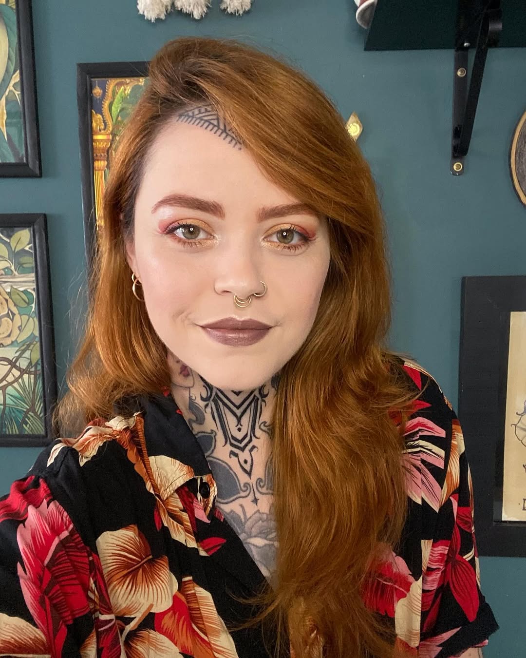 Woman with reddish-brown hair displaying a geometric chest and neck tattoo in black ink with symmetrical line patterns.