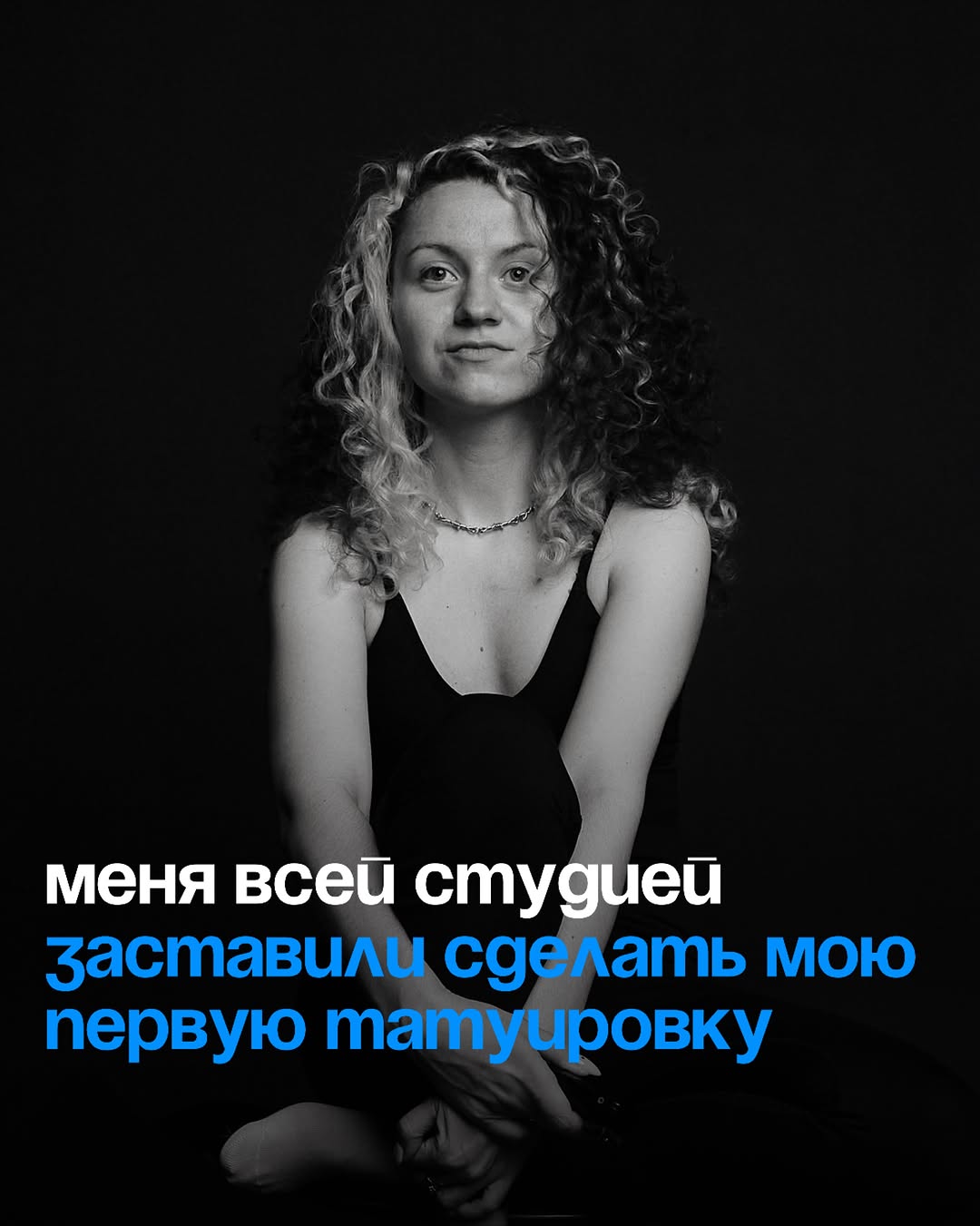 Portrait photograph of a woman with curly hair wearing a delicate chain necklace and dark sleeveless top against black background.