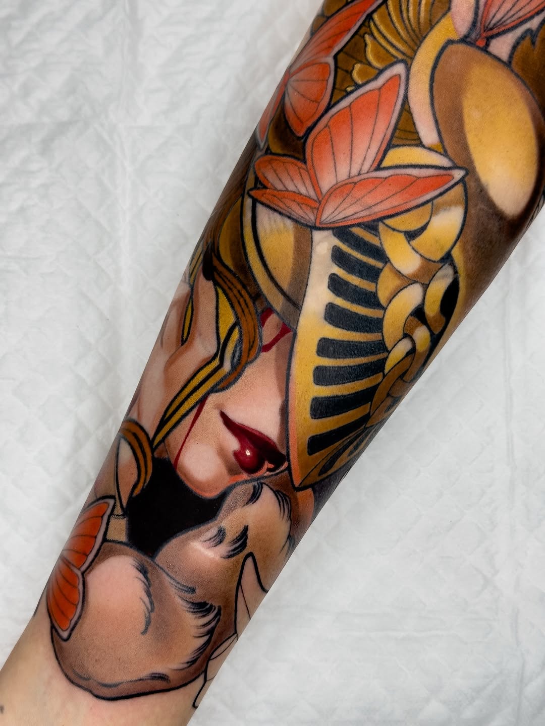 Colorful arm tattoo featuring a woman's face with red lips, gold headdress, orange flowers, and black striped details in yellow and orange tones.