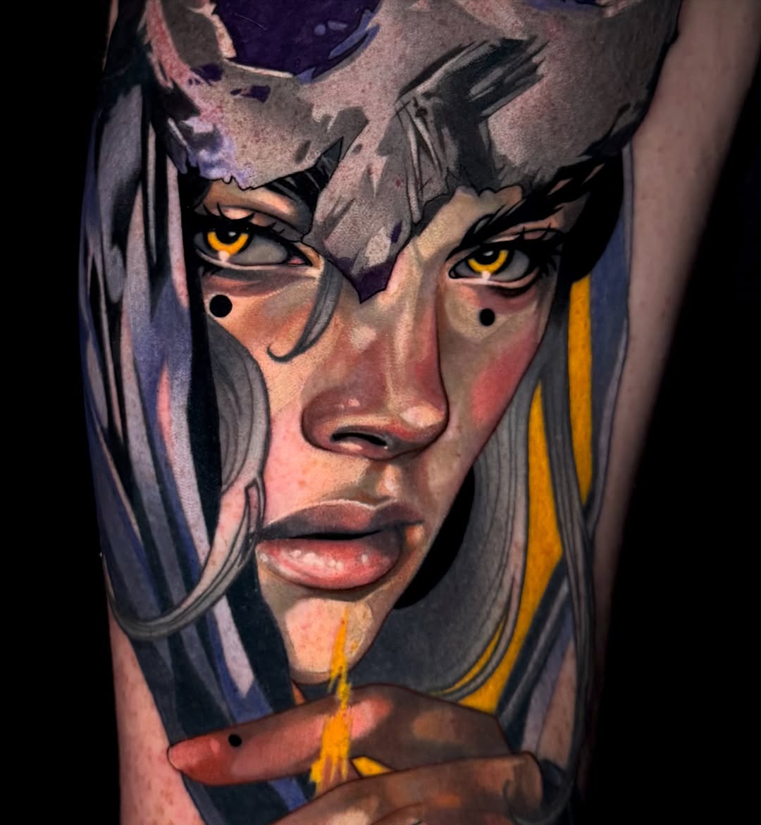Colorful portrait tattoo on upper arm featuring a face with striking yellow eyes, long grey hair, and blue-grey facial markings with yellow and orange accent stripes.