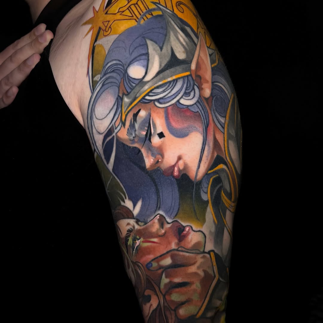 Colorful upper arm tattoo featuring a blue-masked face with gold accents and a smaller portrait below, displaying vibrant blues, golds, and flesh tones.