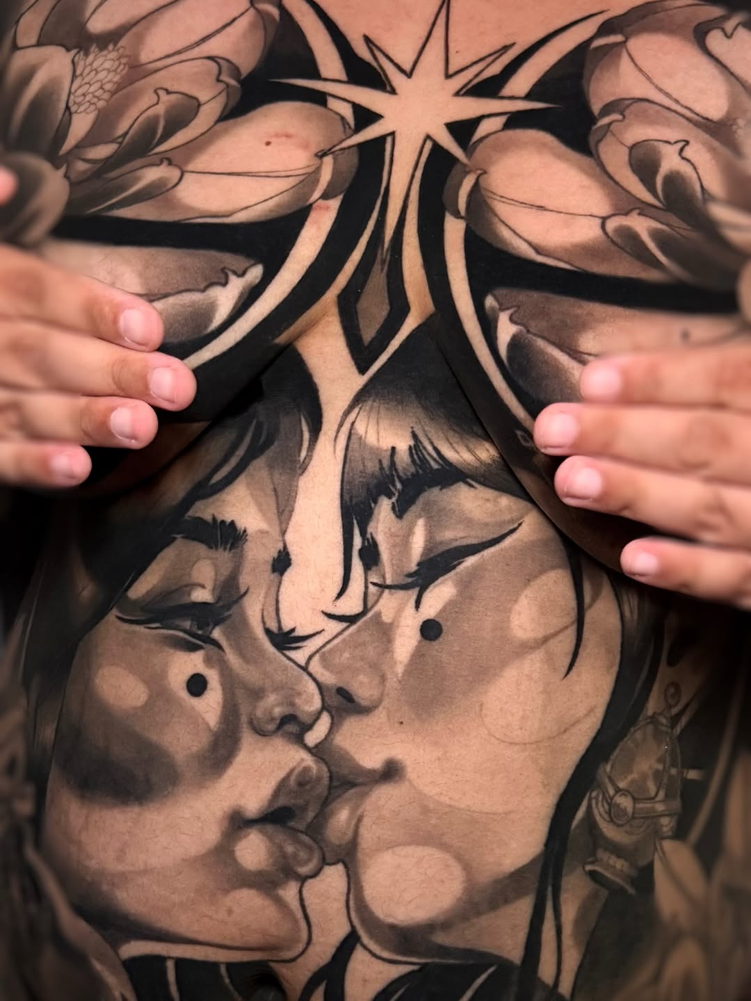 Chest tattoo featuring two facing profiles with closed eyes, flowering vines above, a star, and decorative line work in black and grey ink.