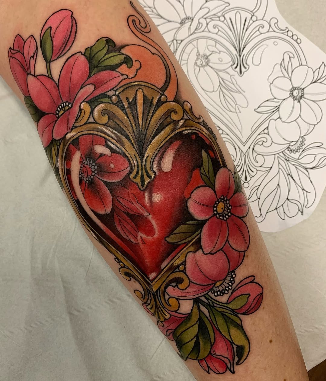 Colorful heart tattoo surrounded by red and pink flowers with ornate gold scrollwork frame on upper arm, displayed next to a sketch design.