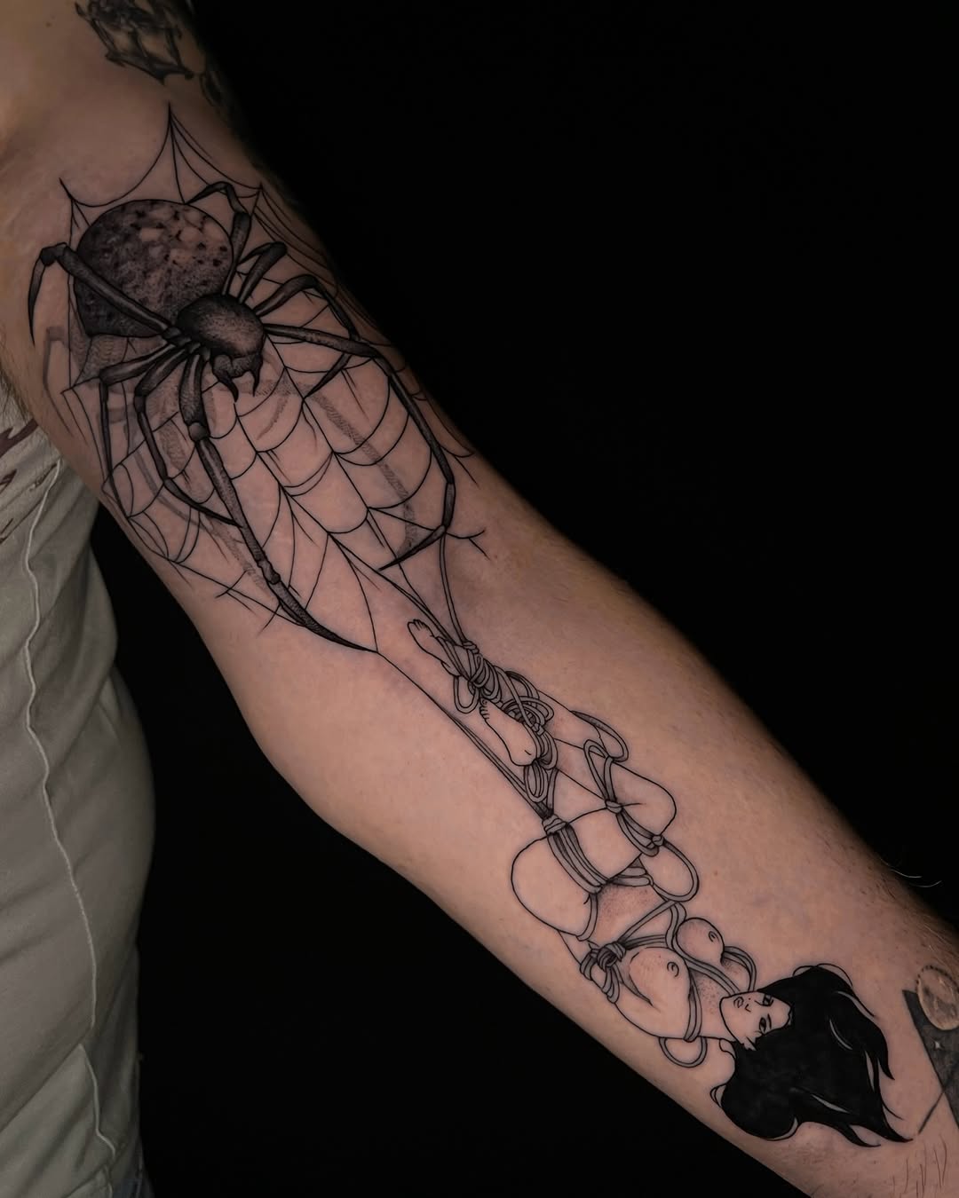 Arm tattoo featuring a detailed spider on a web at the shoulder and a silhouetted figure holding strings extending down the forearm in black ink.
