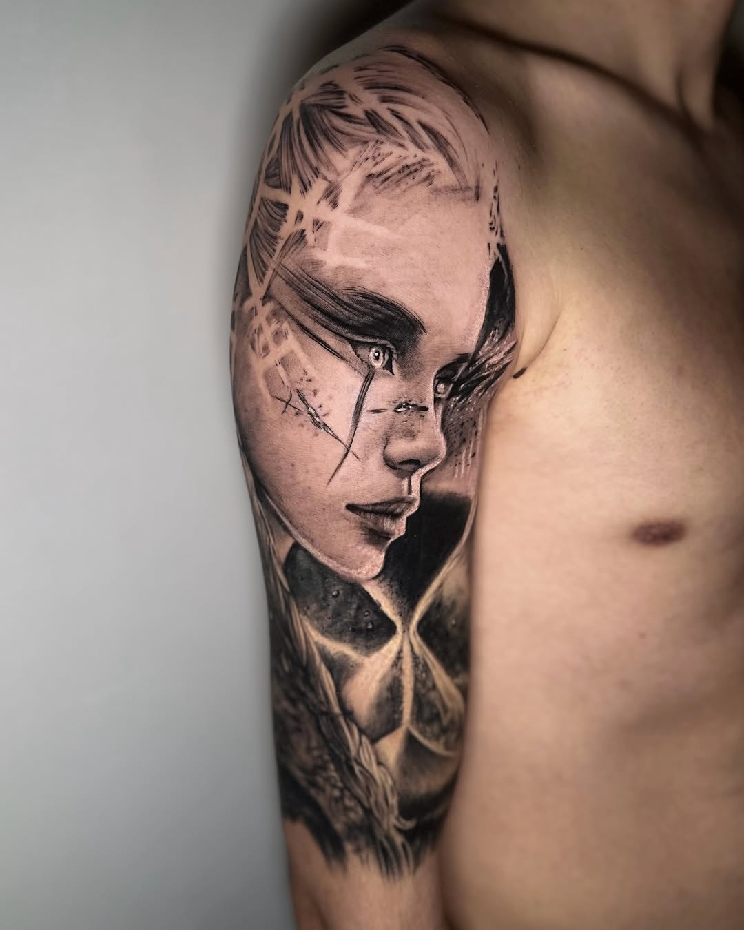 Profile portrait of a woman's face with closed eyes and short hair, tattooed on the upper arm in black and grey ink.