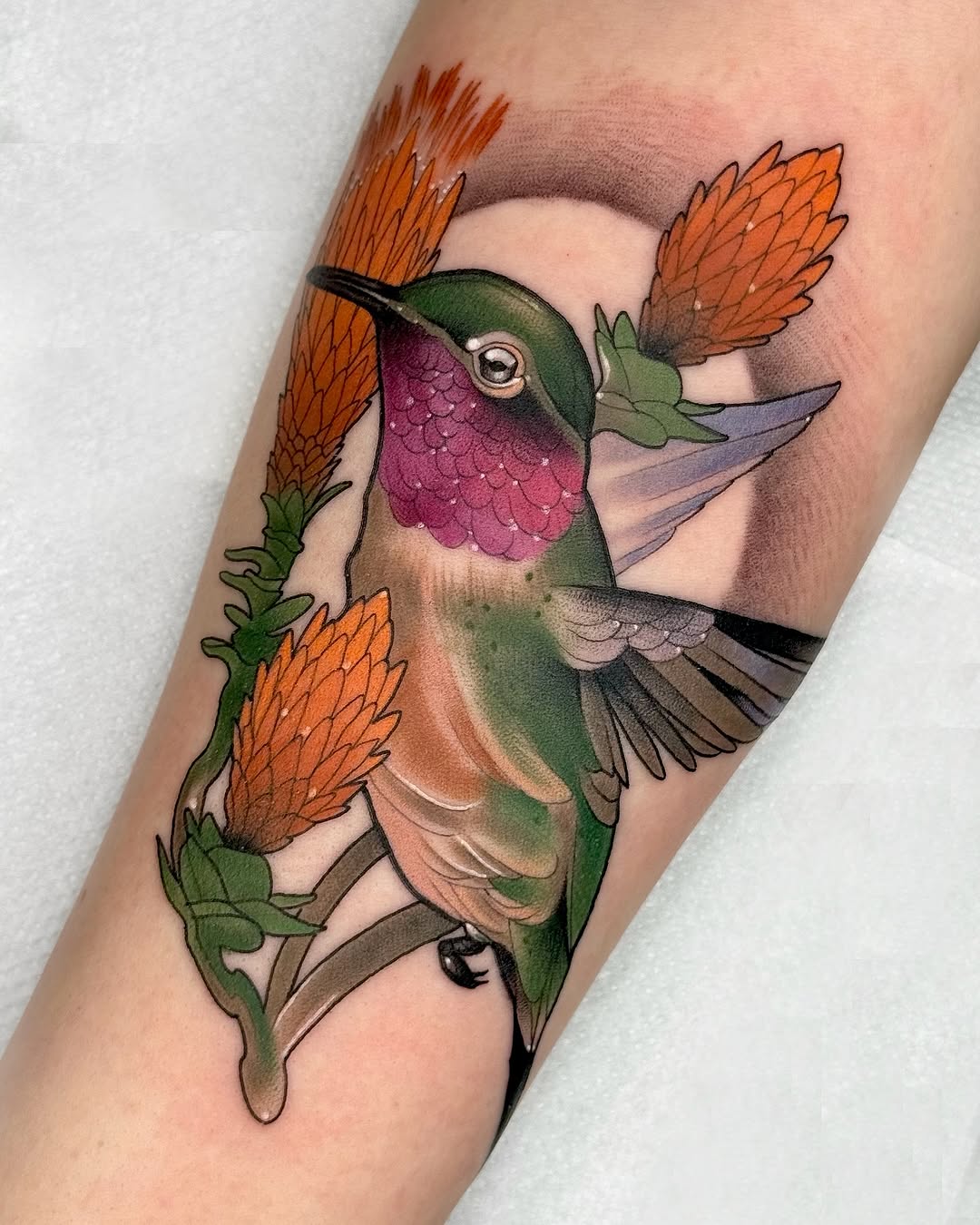 Colorful hummingbird tattoo on forearm with orange flowers, featuring green, purple, and tan plumage against skin.