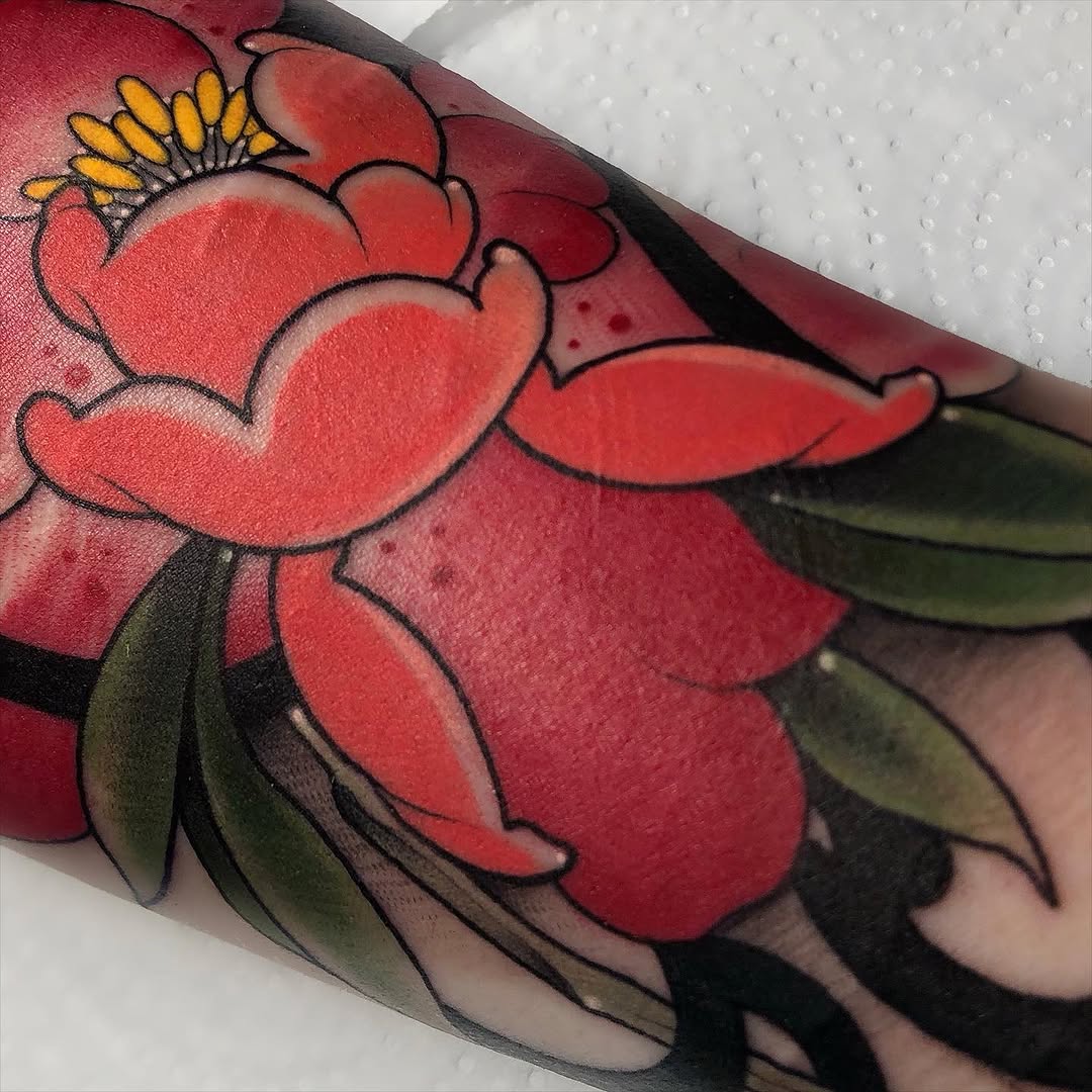 Red flower with yellow stamen and green leaves tattooed on the upper arm, featuring bold black outlines and shaded petals.