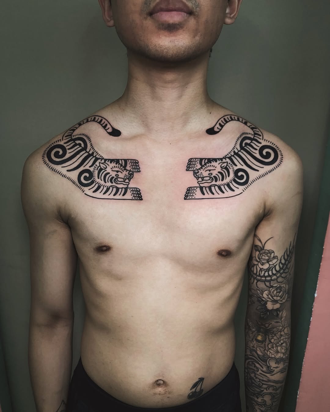 Symmetrical chest tattoo featuring ornate winged mythological creatures with scrollwork and decorative elements in black ink.