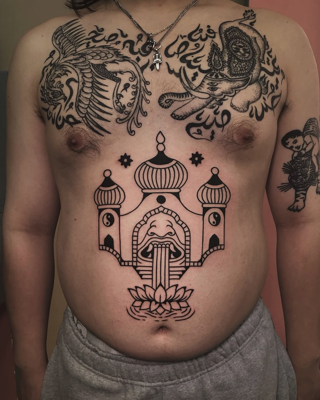 Chest tattoo featuring ornate palace architecture with domed towers, a bearded face with water streams, decorative mythical creatures on shoulders, and lotus flower base in black ink.