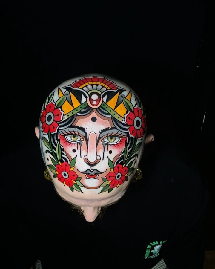 Full-head tattoo featuring a face with red flowers, yellow wings, green leaves, and ornamental designs in red, yellow, green, and black on pale skin.