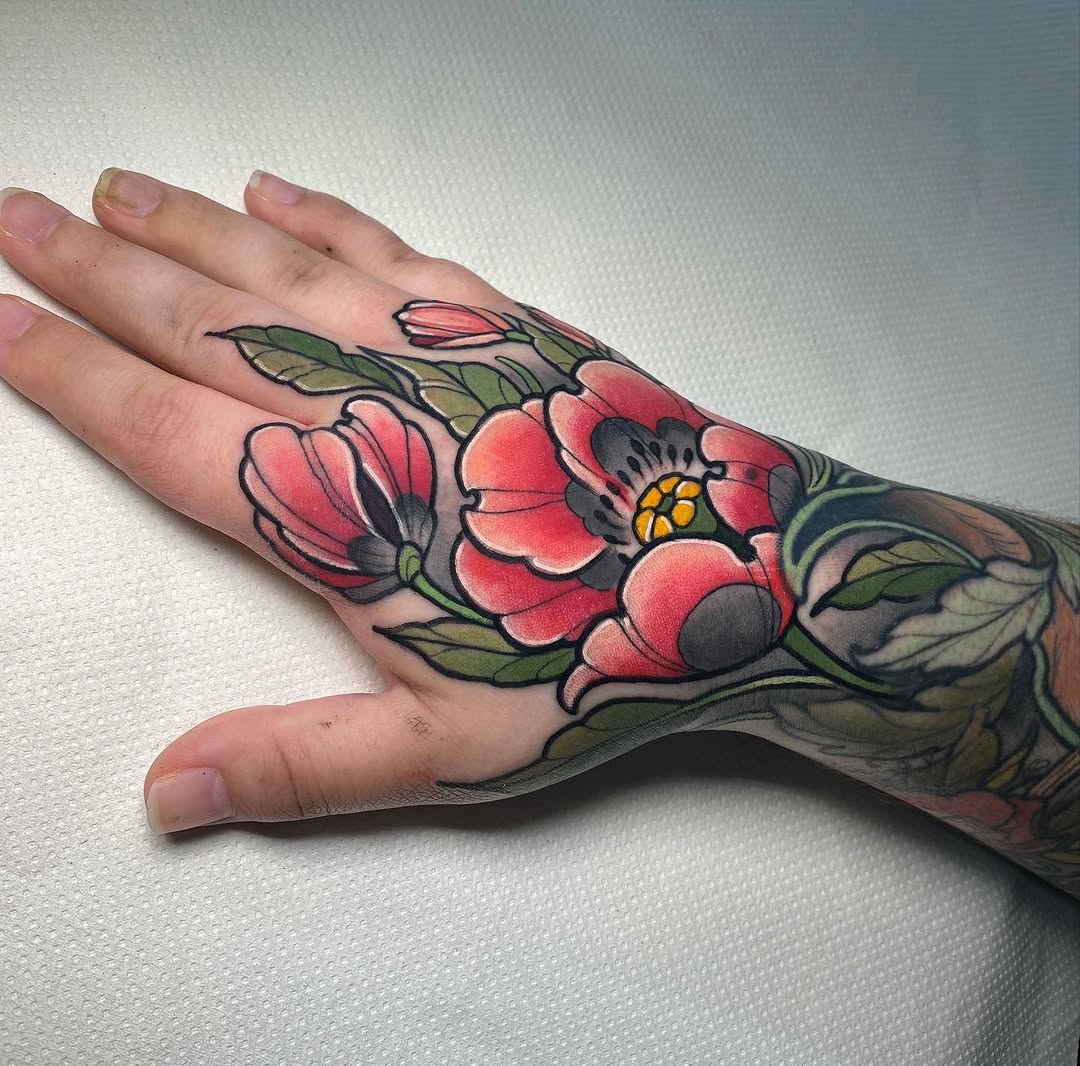 Colorful hand tattoo featuring red and pink flowers with yellow centers, green leaves, and black outlines on the back of a hand and forearm.